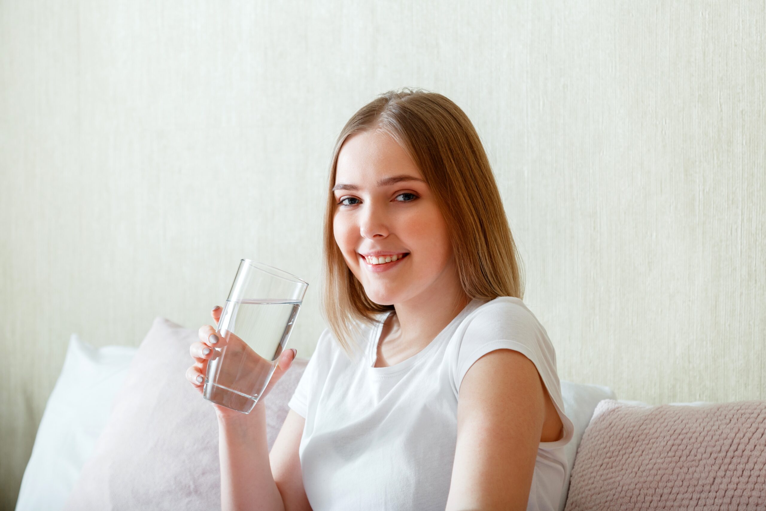 Gülümseyen genç kadın sabah yatak odasında uyandıktan sonra bir bardak saf su içiyor. Mutlu genç kız temiz su içerek vücut sağlığı için su dengesini korur.
