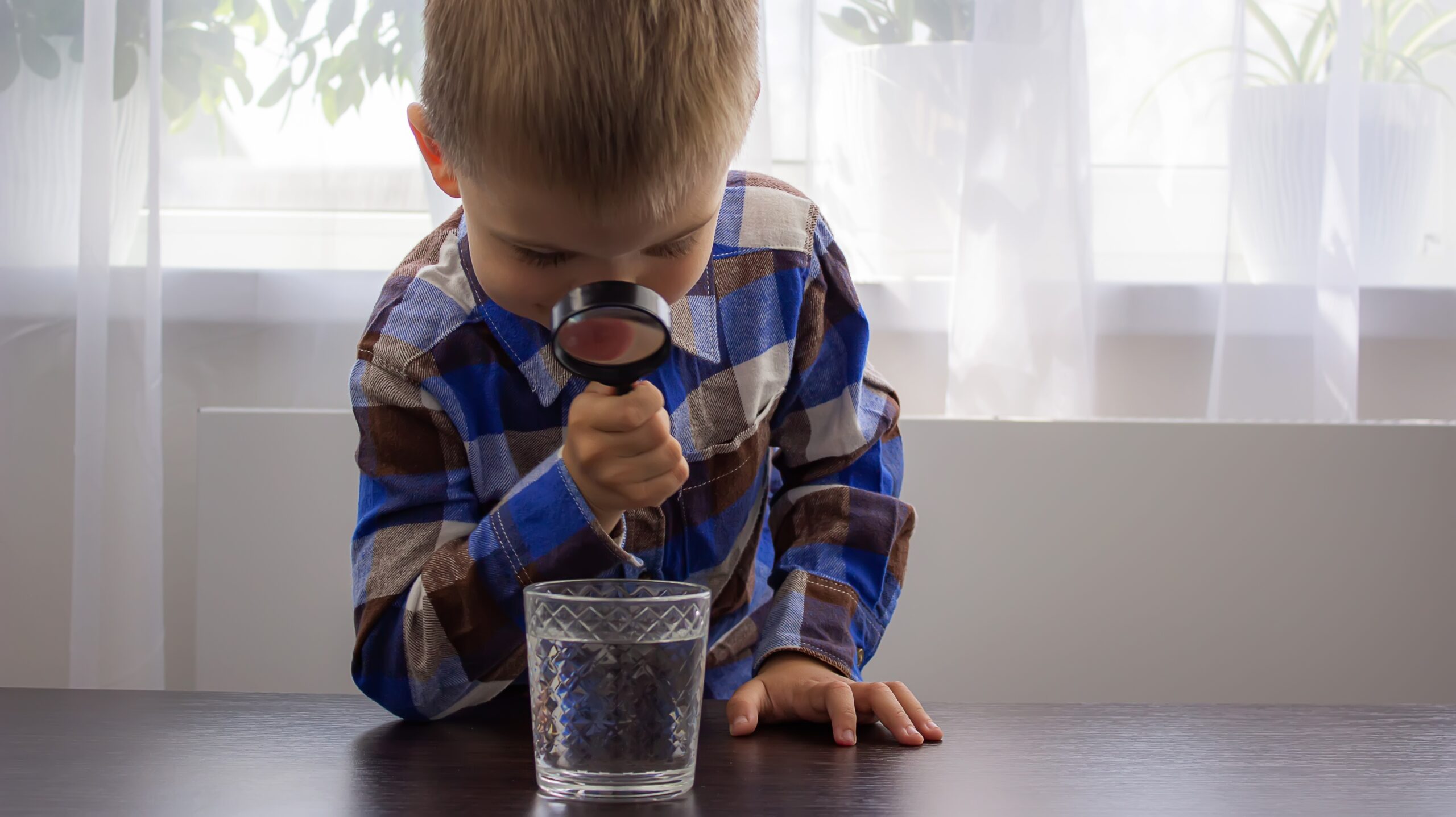 Kareli gömlekli erkek çocuk masa üzerinde duran su bardağındaki suya büyüteç ile bakıyor