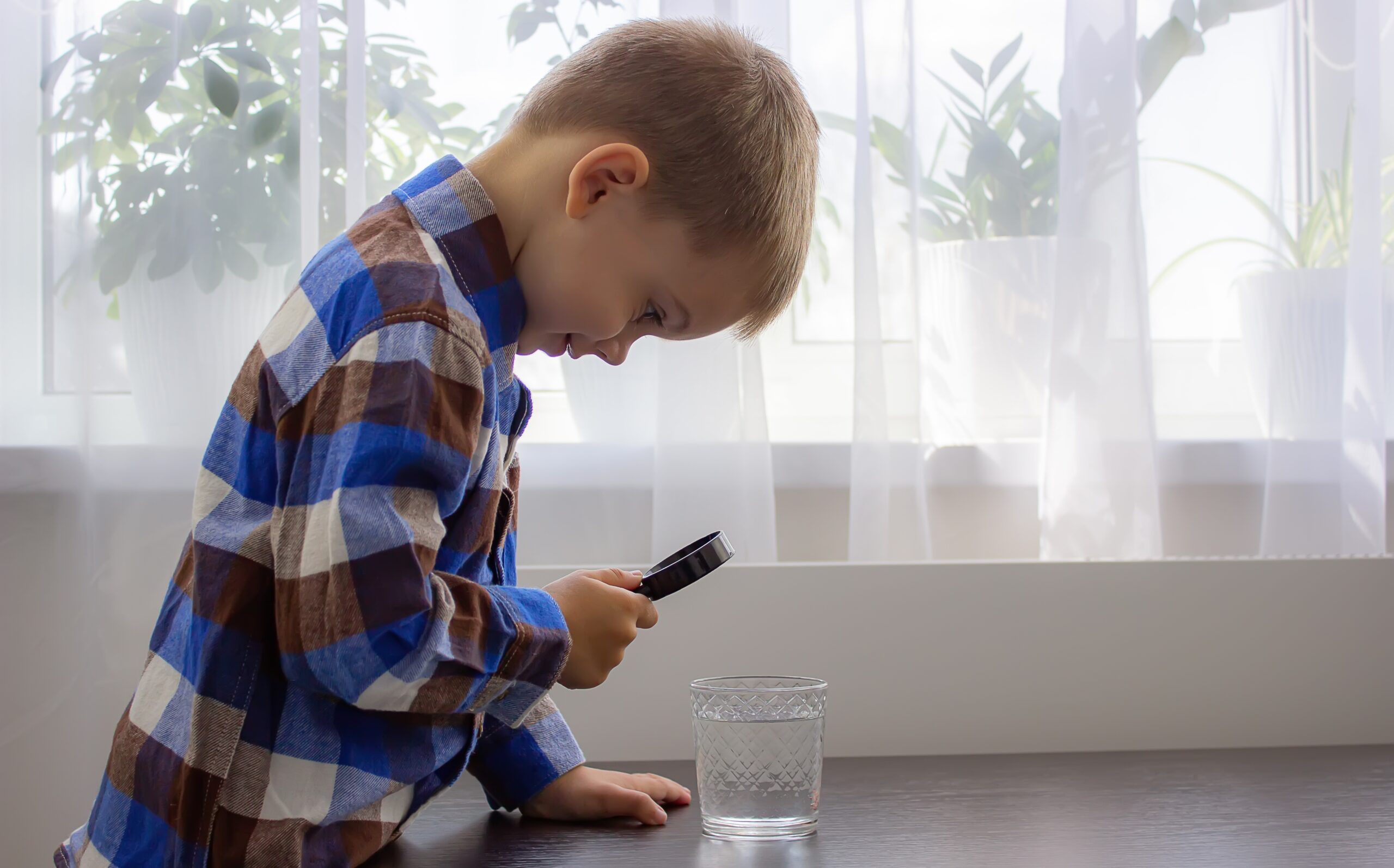 Kareli gömlekli çocuk elinde büyüteç ile su bardağındaki suyu inceliyor