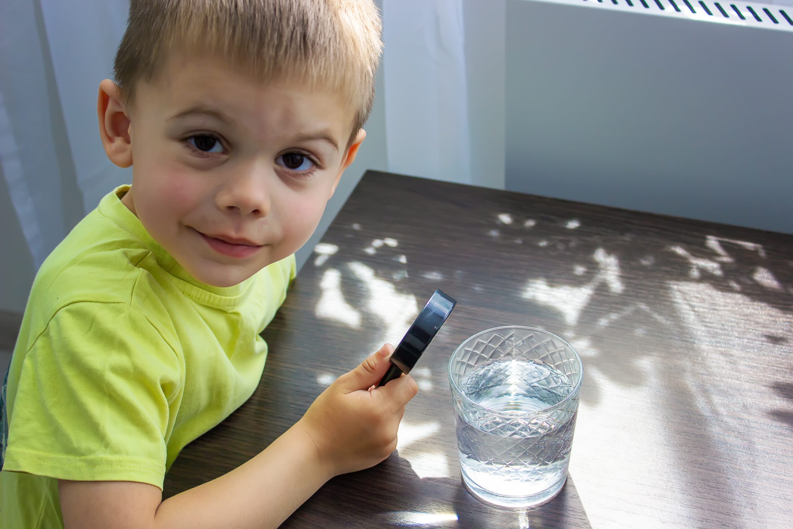 yeşil tişörtlü çocuk elinde büyüteç ile su bardağında ki suyu inceliyor