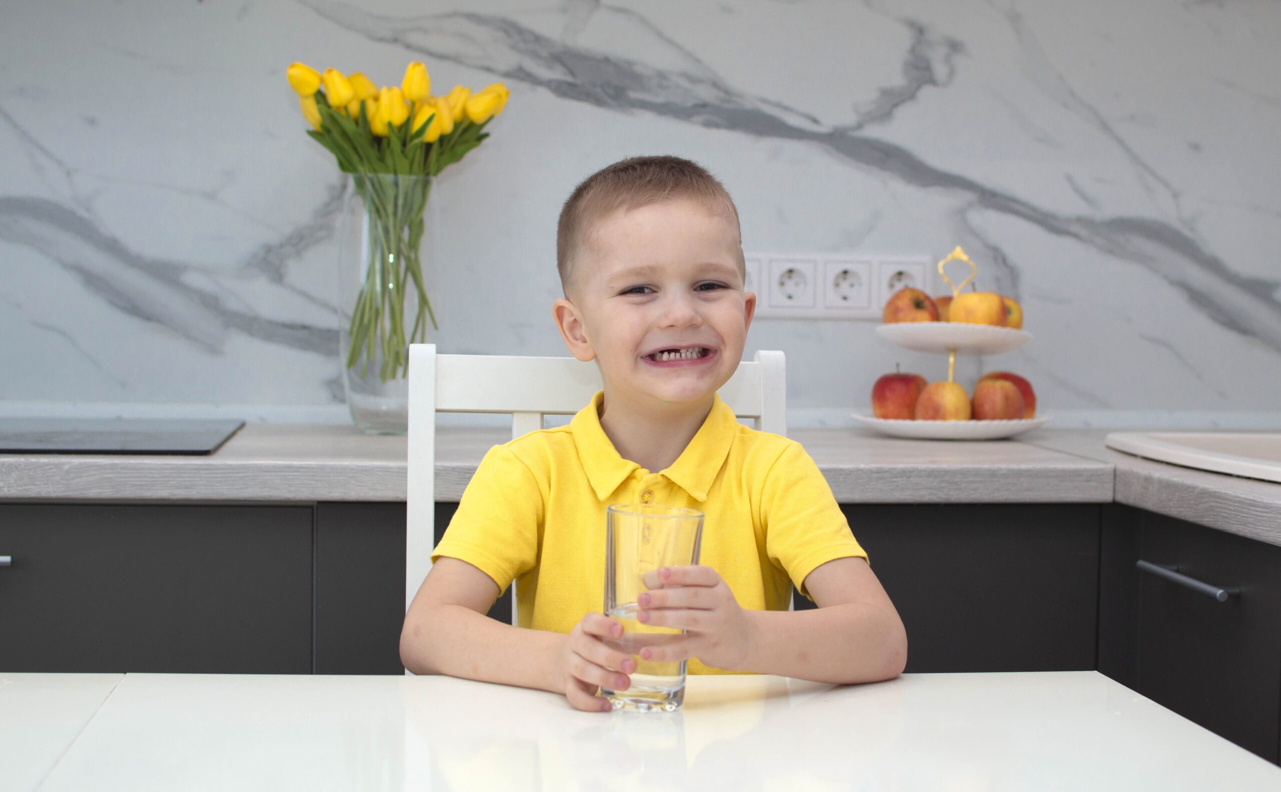 Sarı tişörtlü çocuk evdeki mutfakta su içiyor. Çocuk elinde bir bardak temiz su tutuyor ve gülümsüyor. Su dengesi.