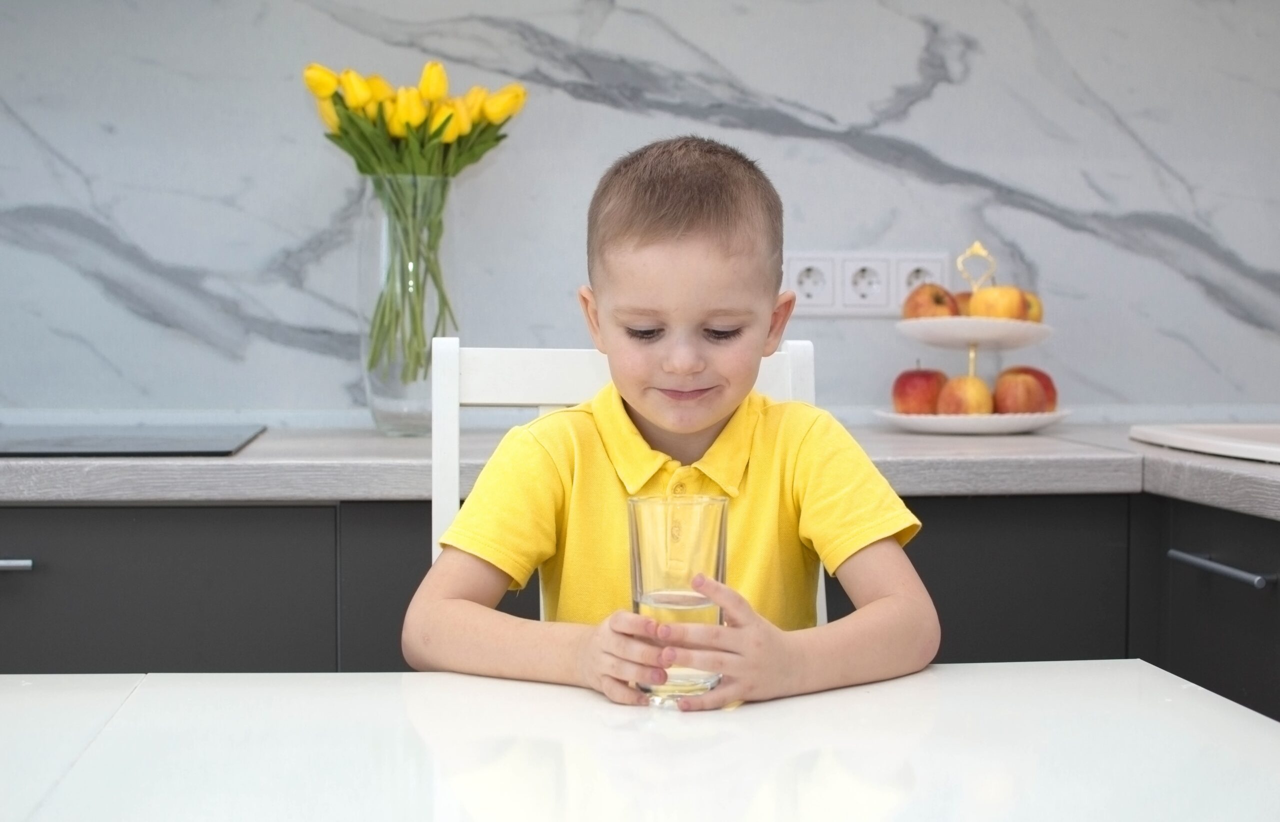 Sarı tişörtlü çocuk evdeki mutfakta su içiyor. Çocuk elinde bir bardak temiz su tutuyor ve gülümsüyor.