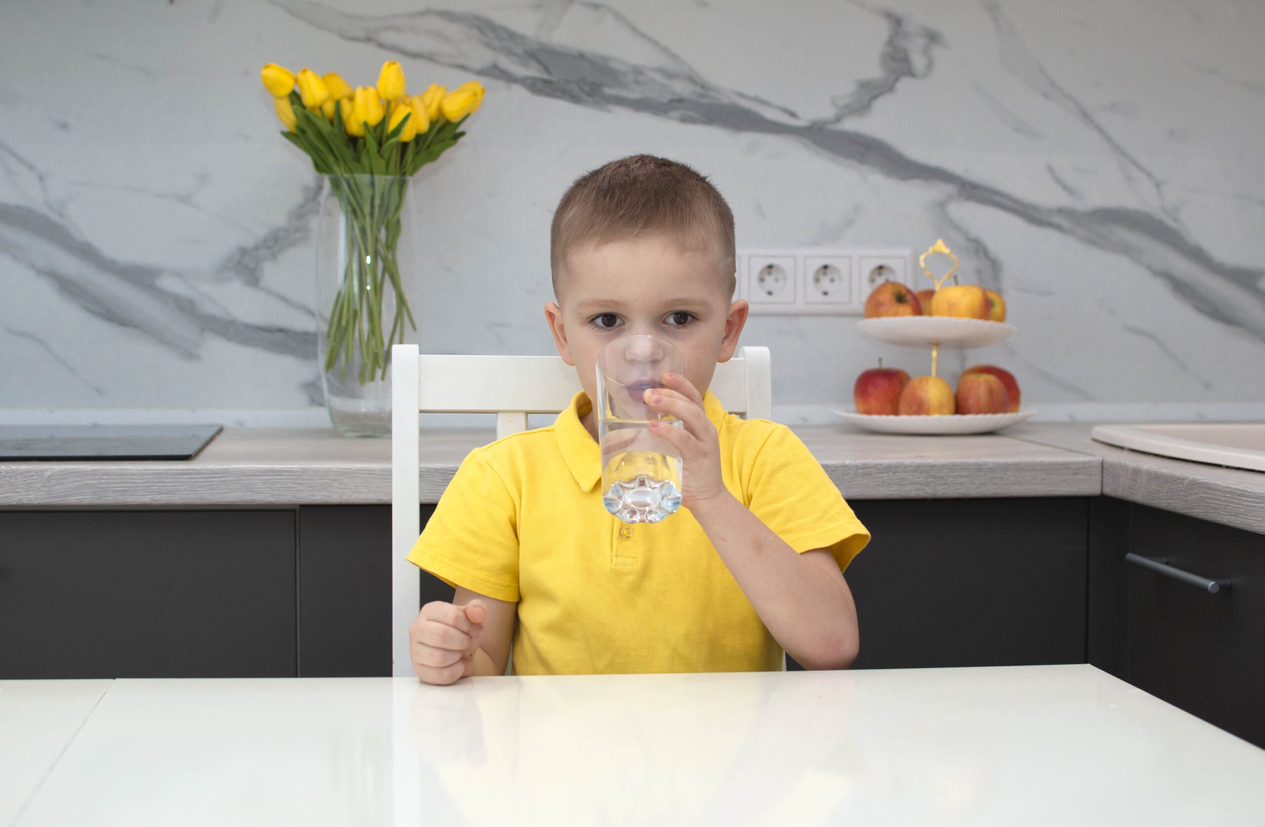 Sarı tişörtlü çocuk evdeki mutfakta su içiyor. Çocuk elinde bir bardak temiz su tutuyor ve gülümsüyor. Su dengesi. Dehidrasyonun önlenmesi