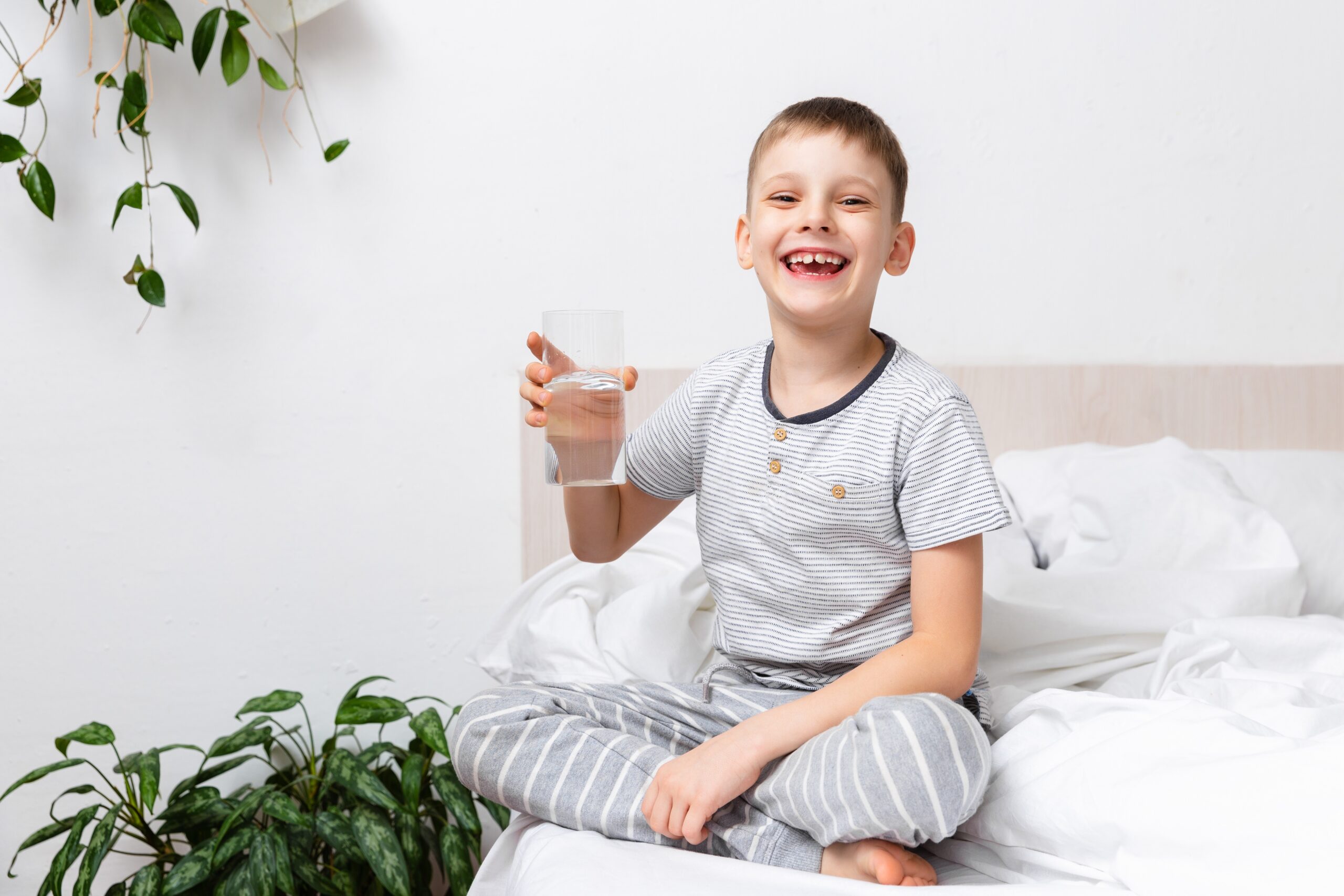 Sağlık kavramı. Mutlu aktif çocuk gülümsüyor, sabahları yatakta oturan bir bardakta taze saf su tutarken. Vücuduna iyi bak. Uyanış Ritüeli