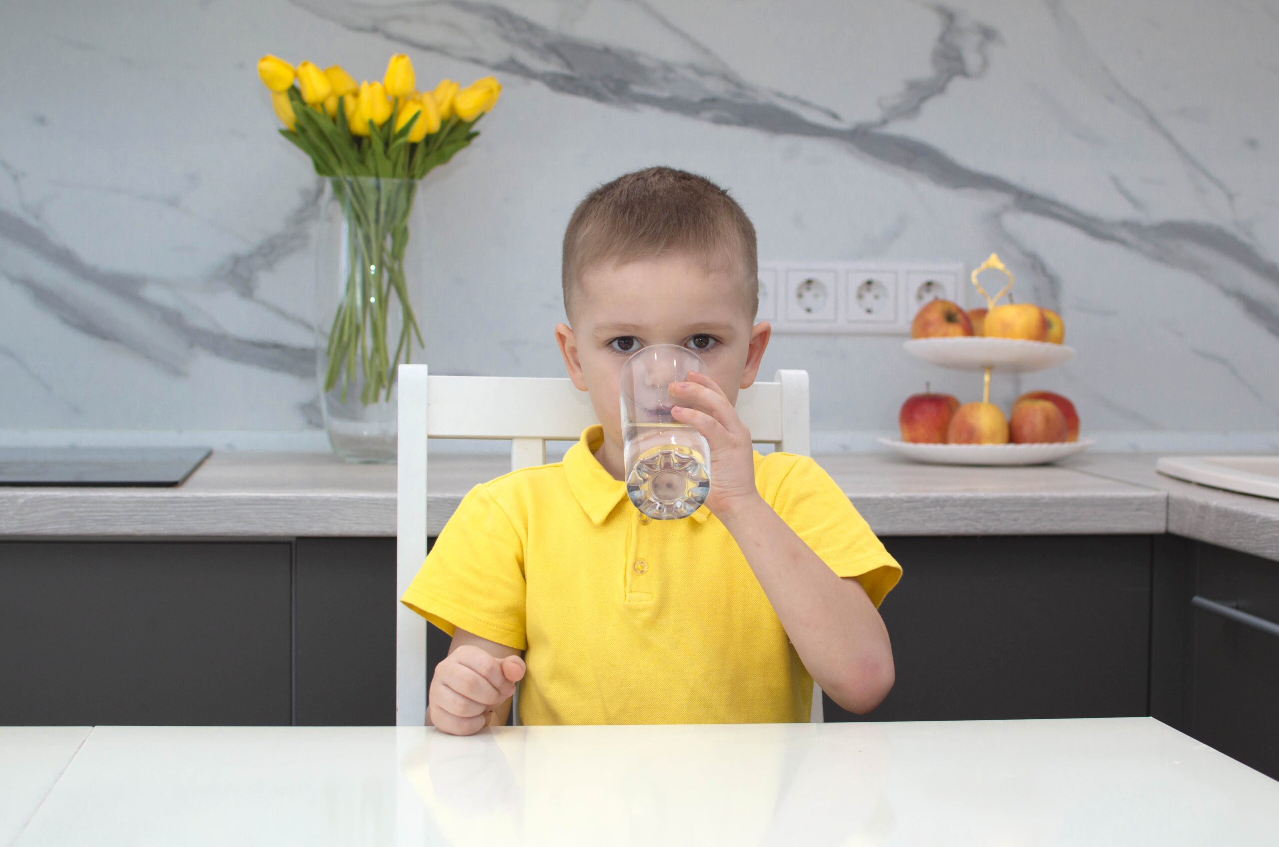 Sarı tişörtlü erkek çocuk evdeki mutfakta su içiyor. Çocuk elinde bir bardak temiz su tutuyor ve gülümsüyor. Su dengesi.